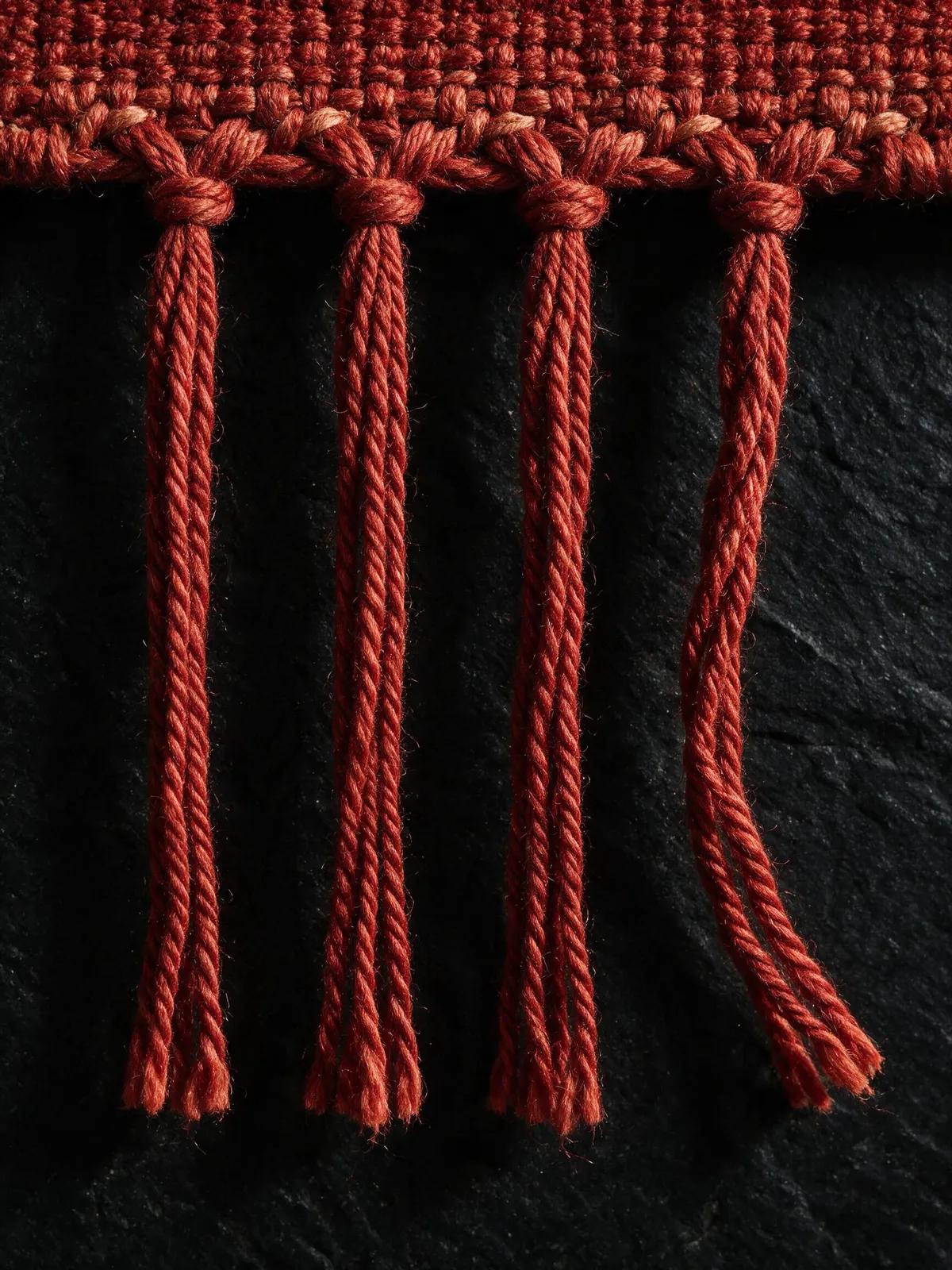 Macro view of madder-red fringe knots against dark basalt stone, raking side light picking out every twist of wool.